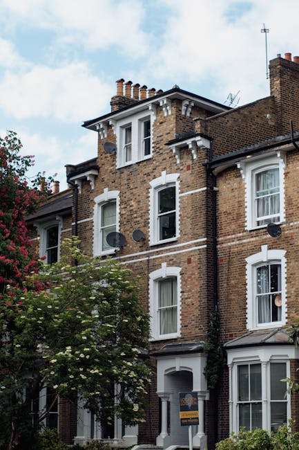 A multi-storey brick residential building on Tufnell Park Road with white-framed windows, some open, and several satellite dishes mounted on the exterior walls. The building features a mansard roof with brick chimneys and decorative white trim around the windows. In the foreground, a flowering tree with green leaves and pink blossoms partially obscures the lower part of the building. The sky above is partly cloudy with patches of blue. This scene relates to house removals and moving services, as part of a typical residential setting in Tufnell Park, London, as highlighted in the Tufnell Park removals guide for N19 flats by Tufnell Park Movers.