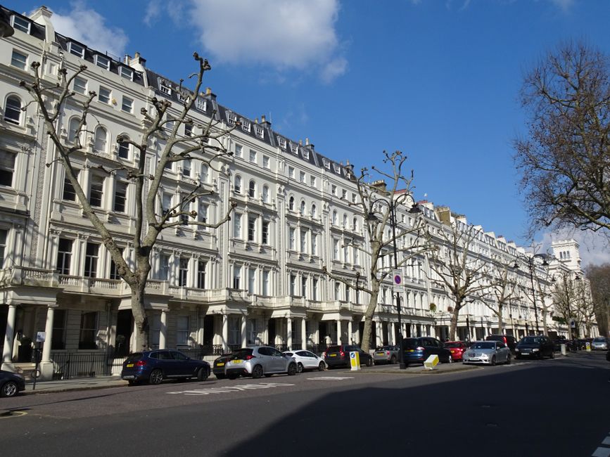 A row of elegant white Victorian-style terraced buildings with large sash windows and ornate architectural details, situated along Tufnell Park Road in N19. The buildings have small balconies and decorative cornices, with a street level featuring black iron railings and entrances. Leafless trees are evenly spaced along the pavement, and multiple parked cars, including black, white, and silver vehicles, line the curb. The scene is captured during daytime with a bright blue sky and some scattered clouds, indicating clear weather. The overall setting reflects a typical residential area suitable for house removals and furniture transport services, with the environment prepared for moving logistics involving loading areas and access points for moving vans, as provided by Tufnell Park Movers.