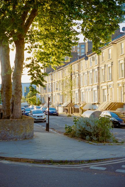 A multi-storey brick residential building on Tufnell Park Road with white-framed windows, some open, and several satellite dishes mounted on the exterior walls. The building features a mansard roof with brick chimneys and decorative white trim around the windows. In the foreground, a flowering tree with green leaves and pink blossoms partially obscures the lower part of the building. The sky above is partly cloudy with patches of blue. This scene relates to house removals and moving services, as part of a typical residential setting in Tufnell Park, London, as highlighted in the Tufnell Park removals guide for N19 flats by Tufnell Park Movers.