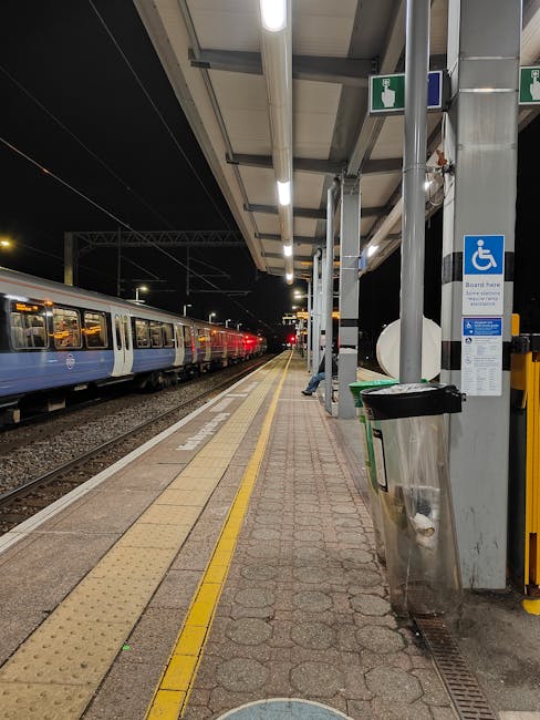 Nighttime view of a London train station platform with a blue and white train waiting at the station. The platform features tactile paving strips along the edge, a covered shelter with metal support b